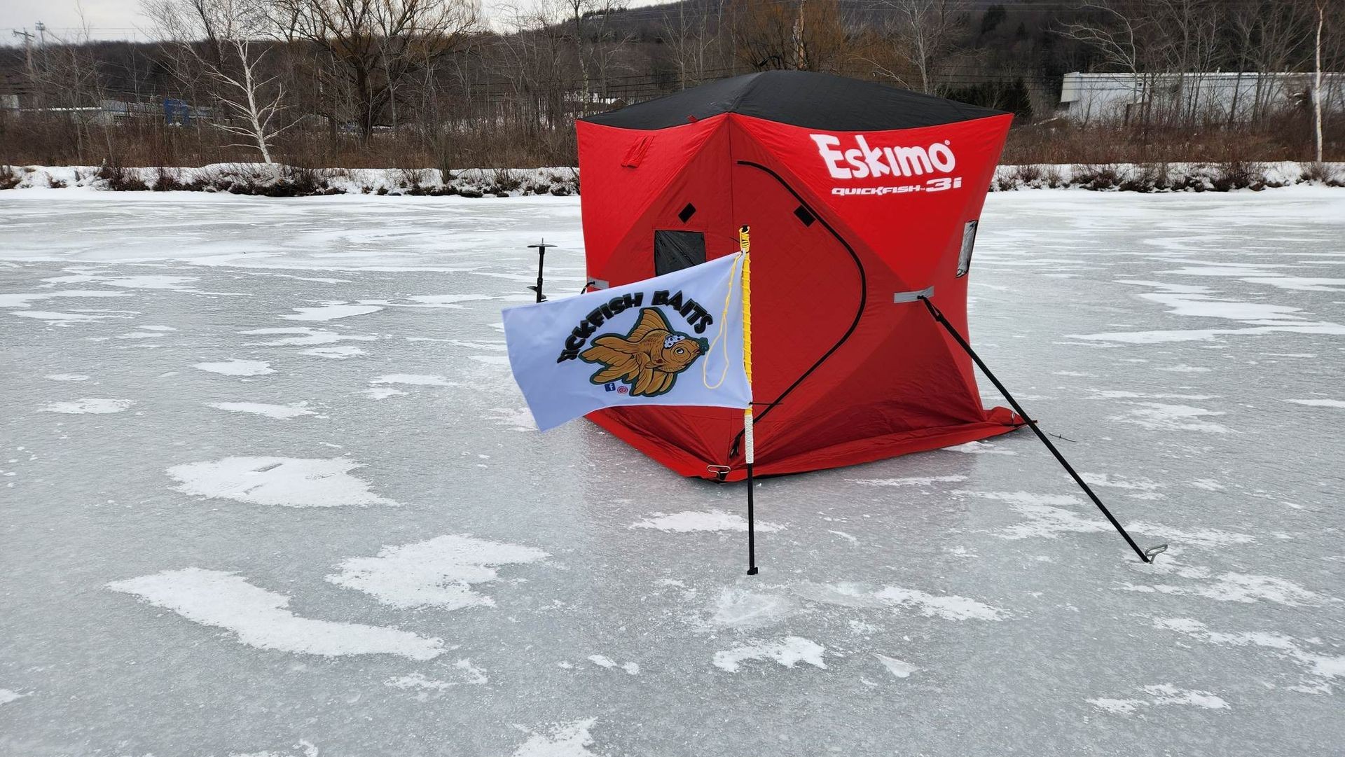 Ice fishing tent located on a frozen pond. There is a flag flying proudly, on it the words "Sickfish Baits" are written in bold black font outlined in gold.